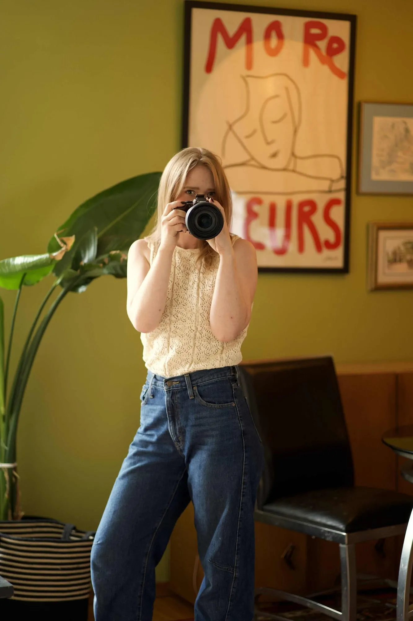 Anna Marishka holding her camera up to her eye, in front of an olive-green wall