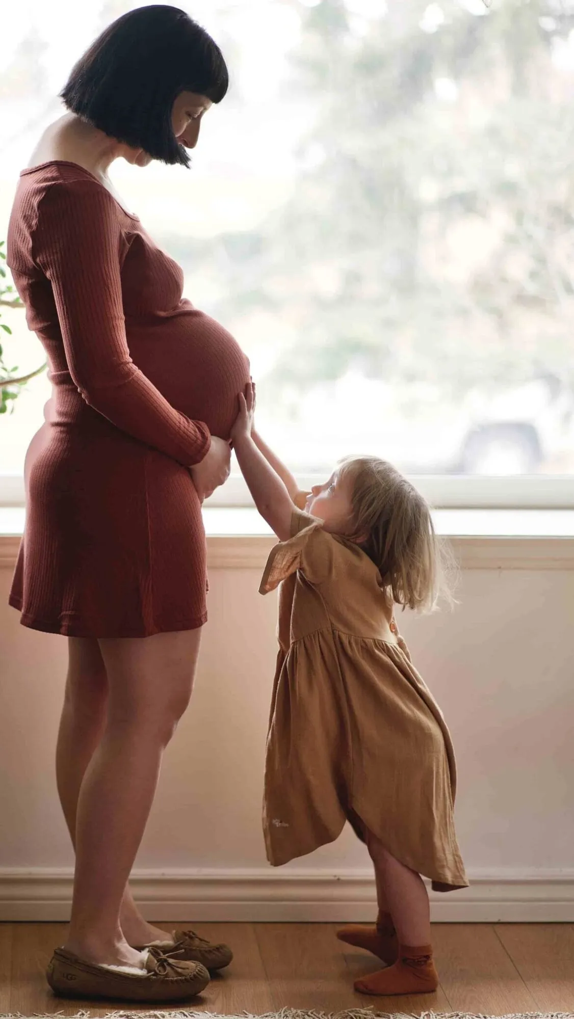 Expectant mother by a bright window as her toddler reaches up to her bump